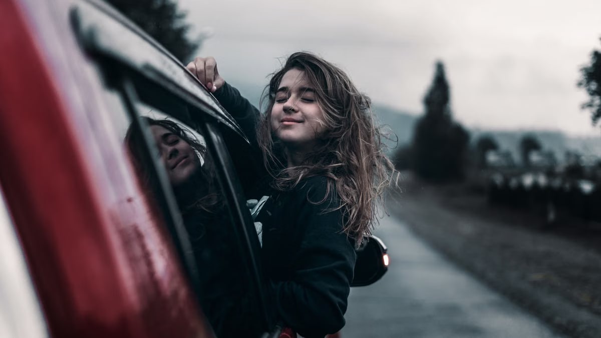 Contented young female with brown hair fluttering in wind leaning out of window of moving car in countryside
